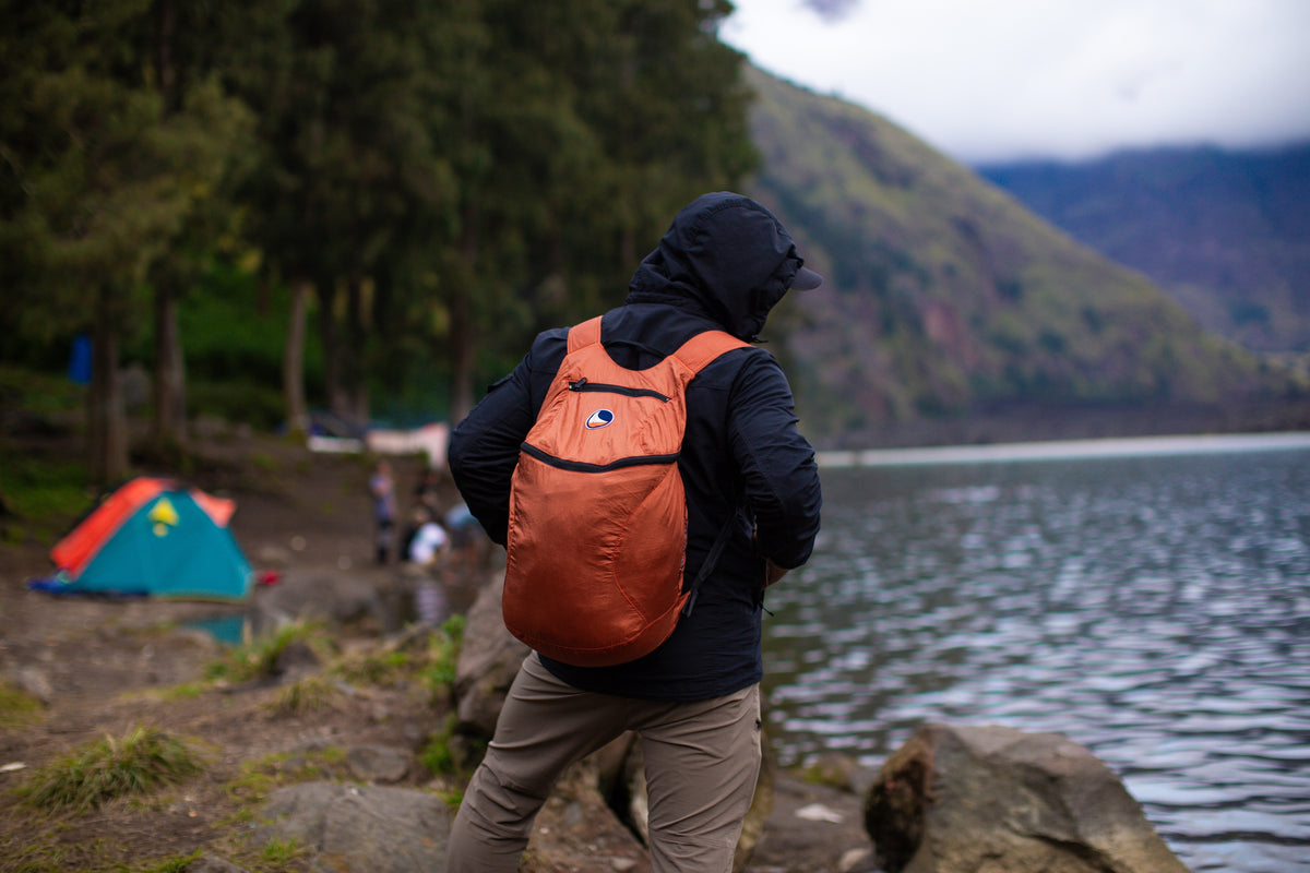 Person hiking by a mountain lake carrying a Copper Backpack Plus 25L, with tents in the background