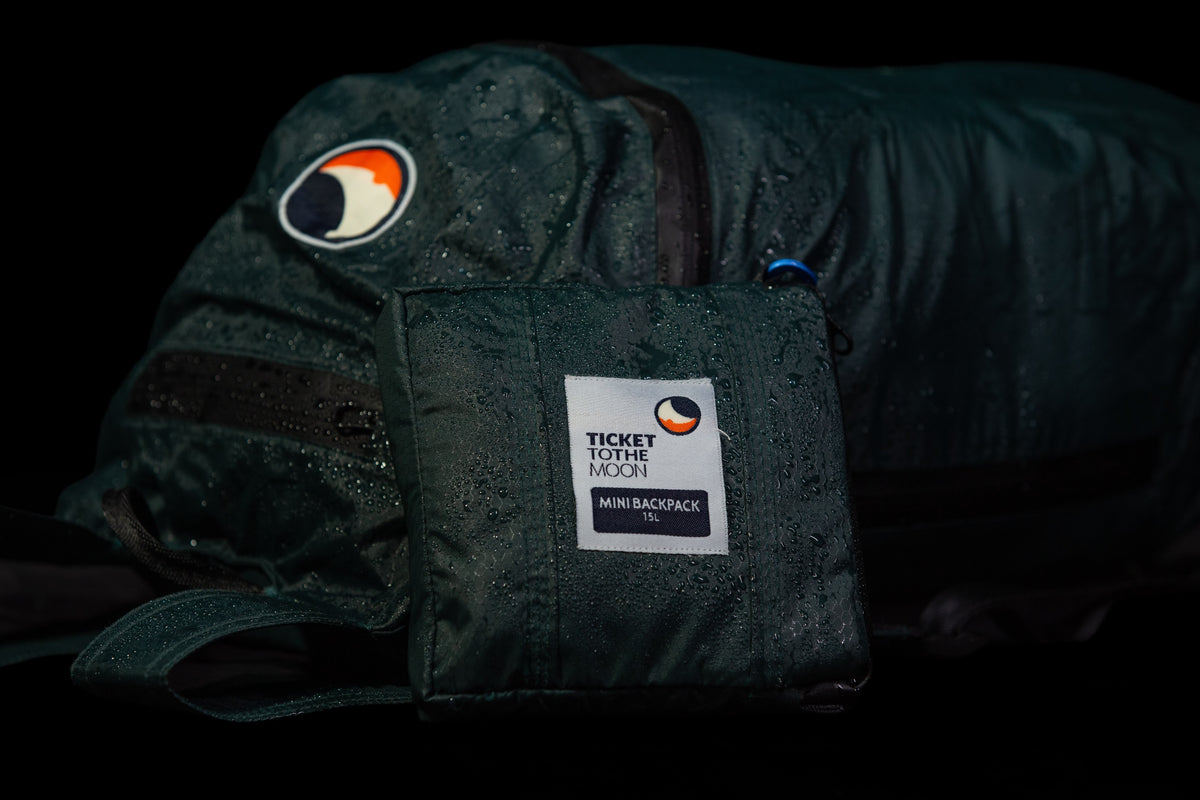 雨粒に覆われたグリーンのバックパックの前に置かれた、専用ポーチに折りたたんだ Mini Backpack 15L